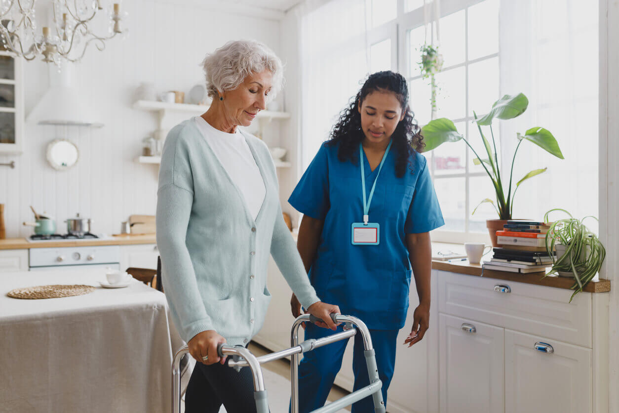 A skilled nurse is helping an older woman with grey hair in a cardigan to use a walker during a care-at-home session.